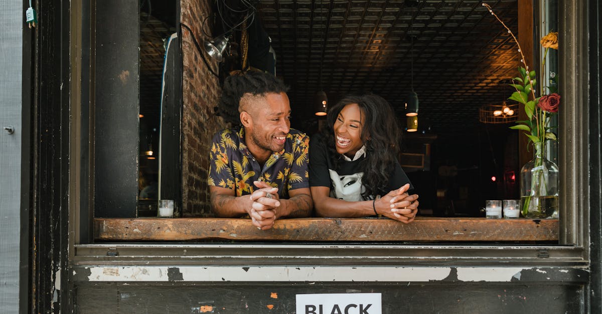 Two joyful business owners lean on a window sill with a 'Black Businesses Matter' sign visible.