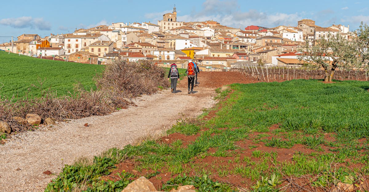 Two pilgrims with backpacks walk towards the picturesque village of Cirauqui, Spain, on a sunny day.