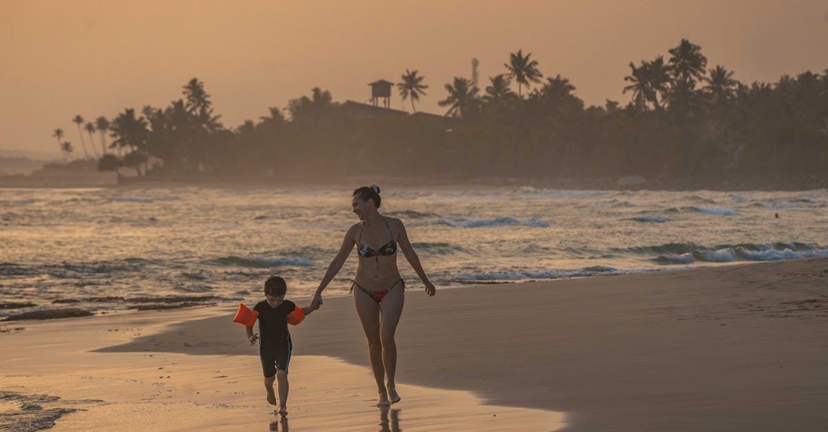 A woman and child holding hands while walking on a scenic beach during sunset.