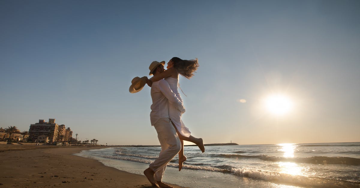 A couple in white attire joyfully embracing on a sunlit beach with a distant city view.