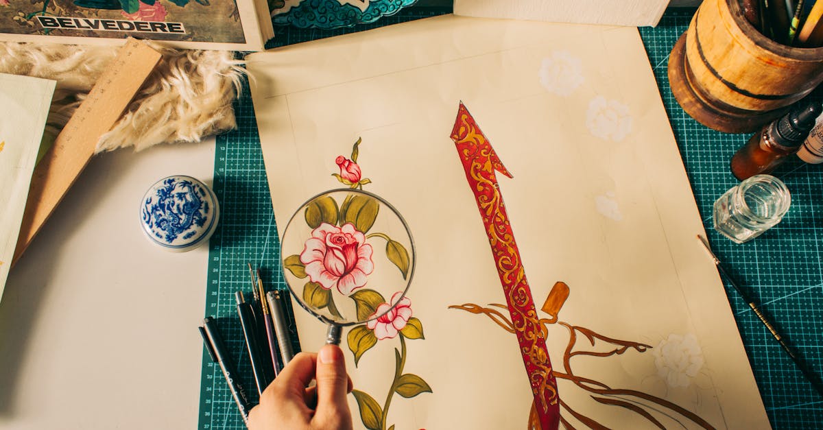 An artistic design on paper with detailed floral motifs, tools, and magnifying glass on a workspace.