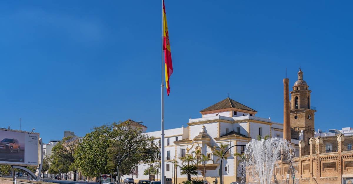 Scenic view of a plaza in Cádiz with a prominent Spanish flag, a fountain, and historic architecture.