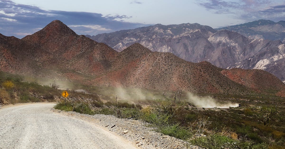 Curved dirt road amidst arid landscape with towering mountains under a blue sky.