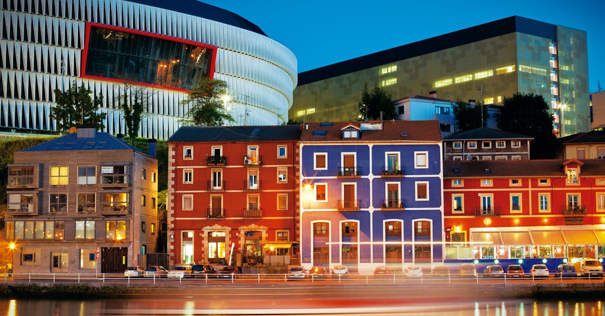Colorful riverside buildings and San Mamés Stadium in Bilbao illuminated against the night sky.