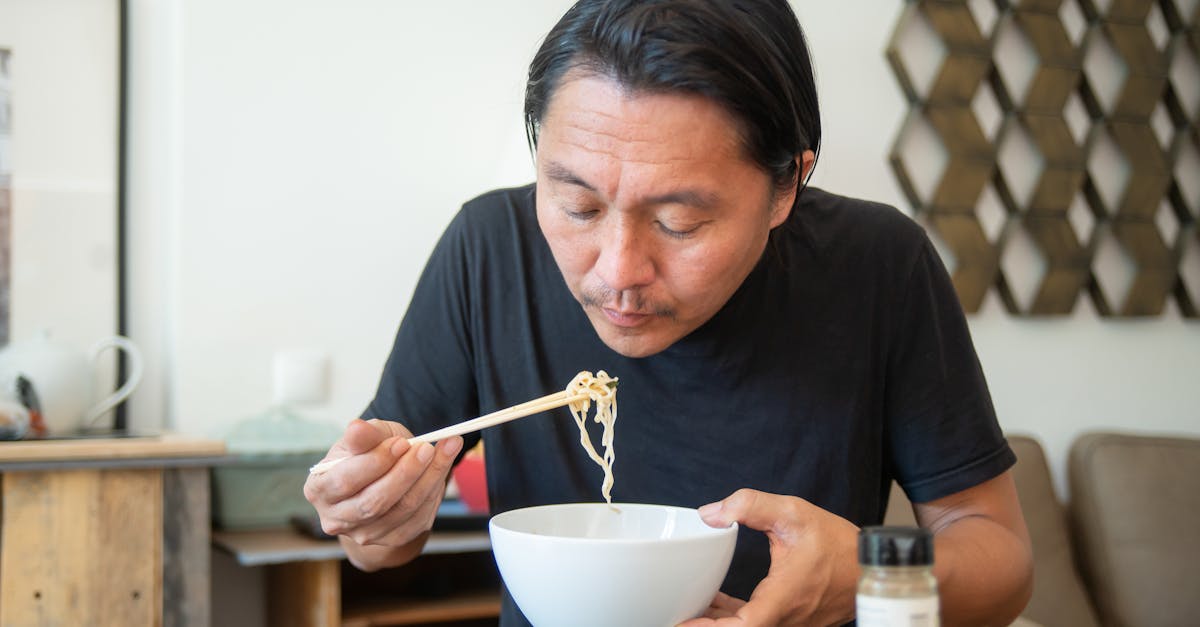 An Asian man eats noodles with chopsticks in a cozy indoor setting, showcasing a homey atmosphere.