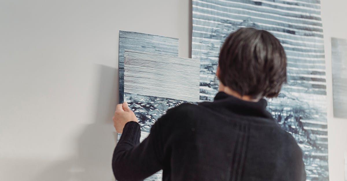 A woman in a dark sweater arranging abstract paintings on a wall, showcasing creative expression.