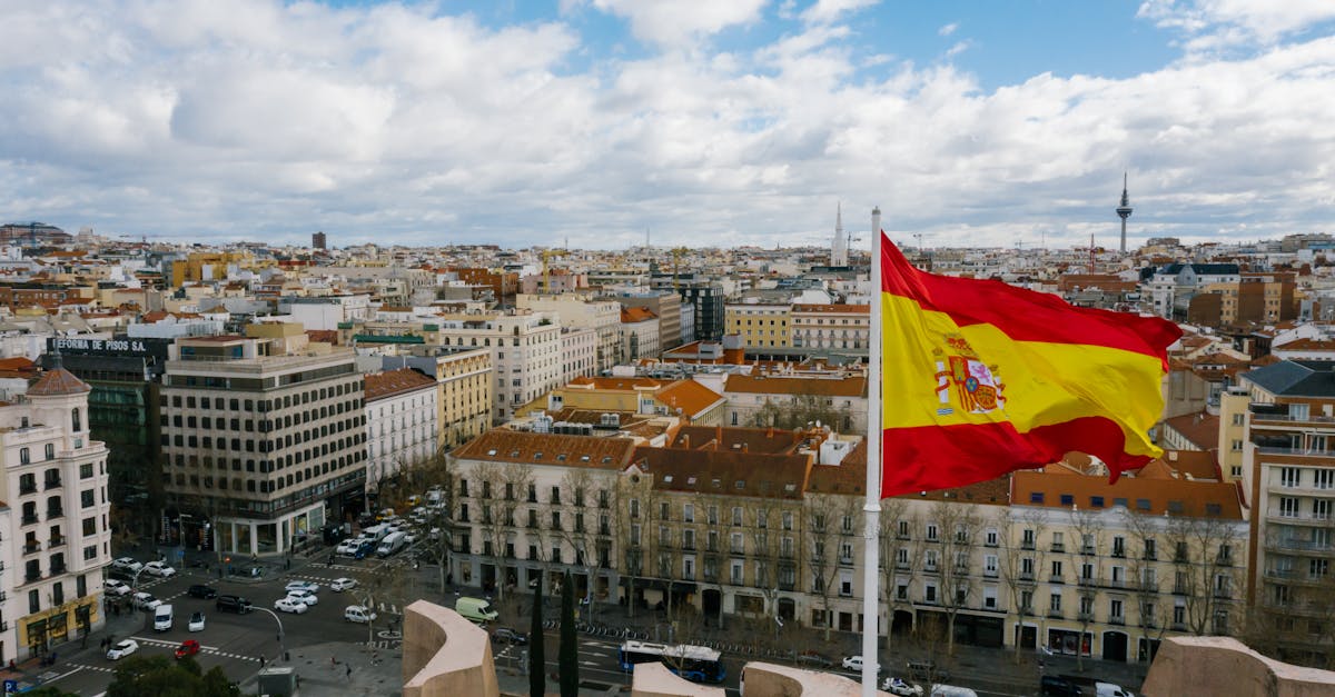 Drone view of Spanish city with aged buildings and national flag under cloudy blue sky