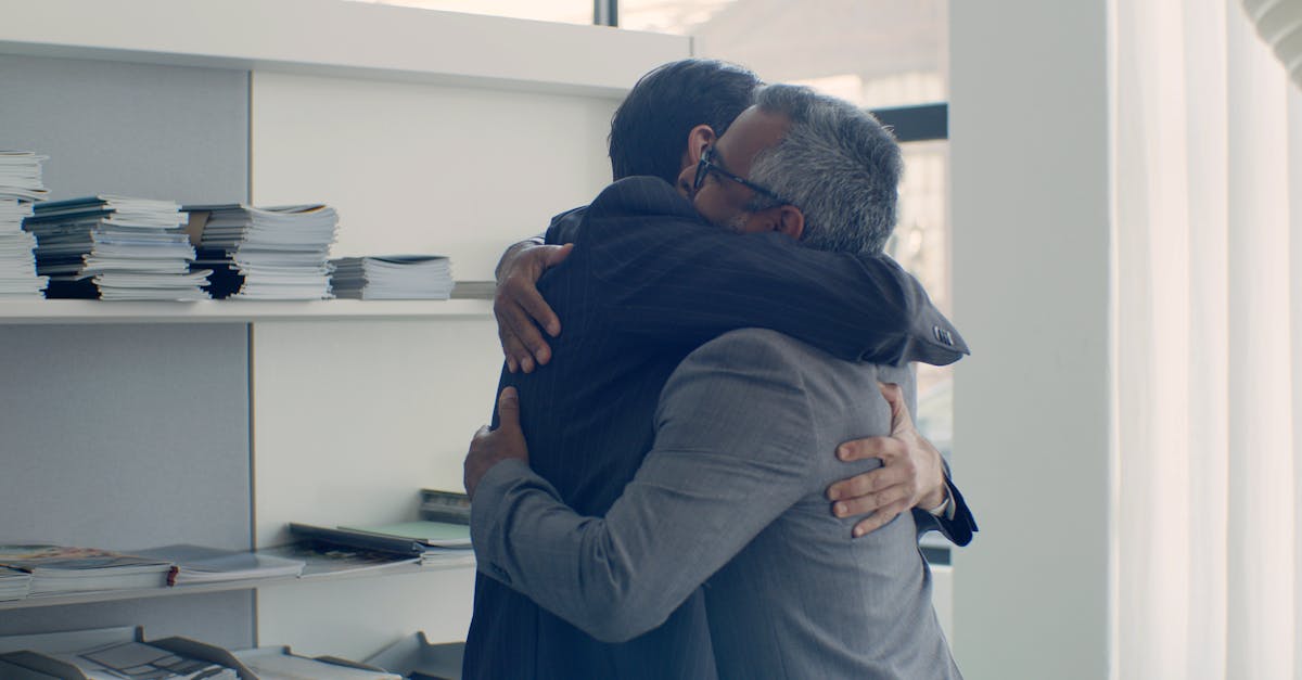 Two businessmen in suits share a warm hug in a sunlit office.